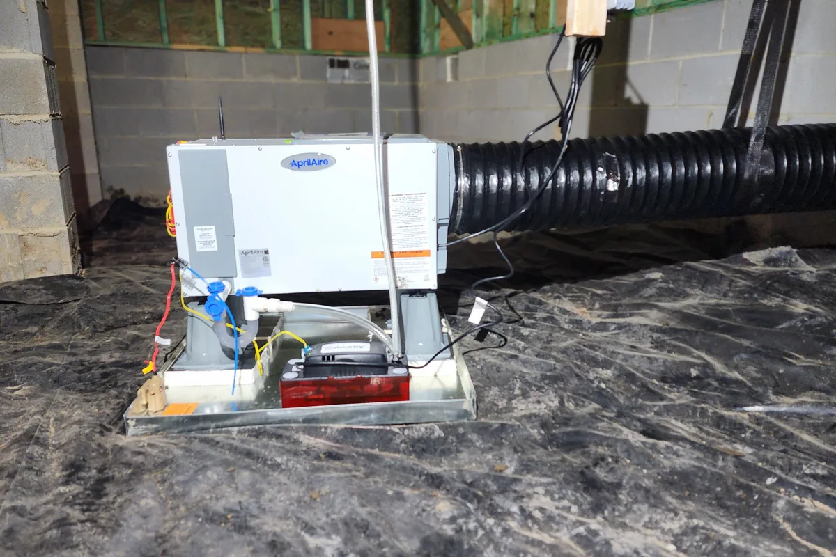 AprilAire crawl space dehumidifier in a drain pan with a condensate pump over a black ground vapor barrier, with unlined block foundation walls and piers, in a Triangle NC crawl space
