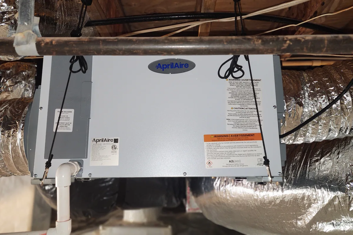 AprilAire whole-house dehumidifier suspended in ductwork with flex duct connections on both sides in a Wake County NC attic